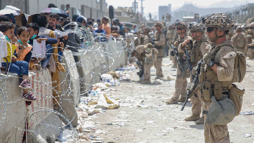 Afghans (left) stand in front of a wall, barbed wire and armed US soldiers