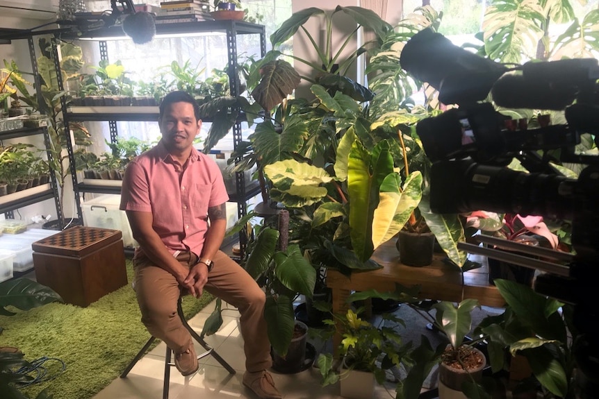 Dexter Burgos sits in a room surrounded by tropical plants while a camera film him.