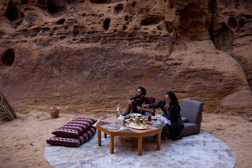 A man and a woman sit at a table on the desert sands.