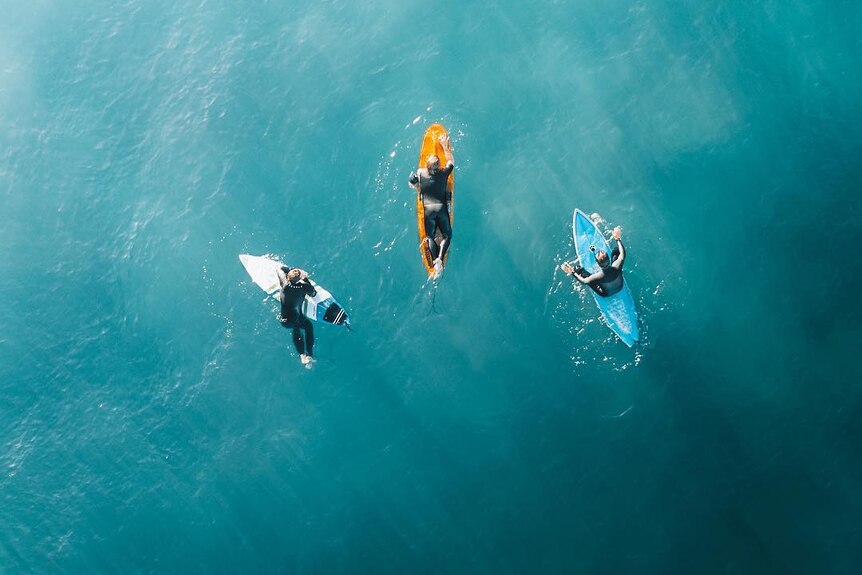 A drone photo of three people sitting on their surfboards.