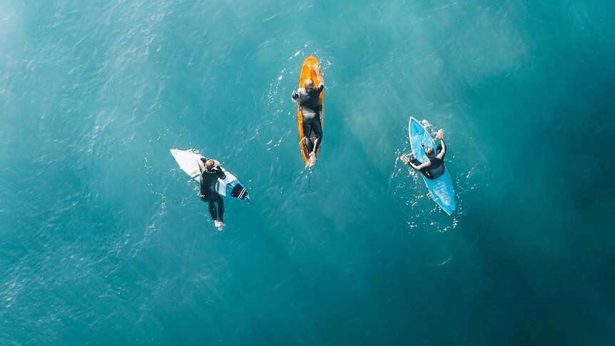 A drone photo of three people sitting on their surfboards.