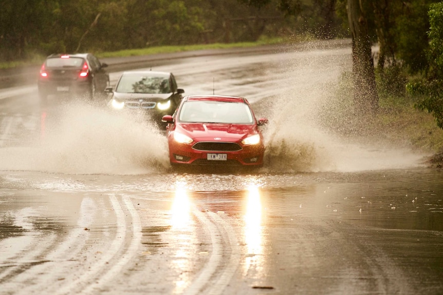 A red car driving through a huge puddle with its headlights on.