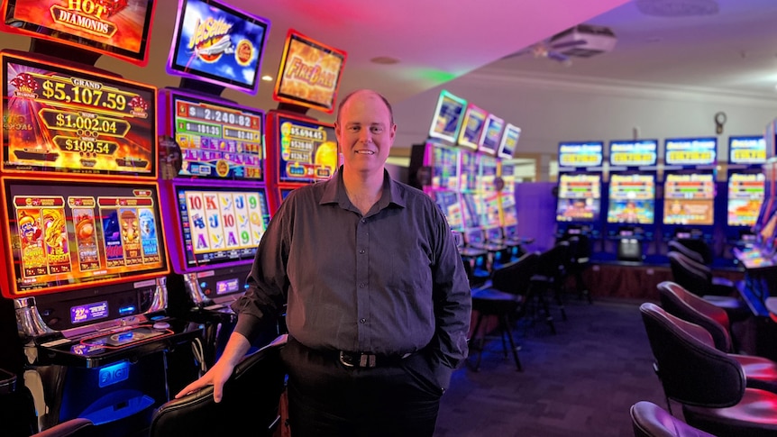 A balding man in a shirt standing in front of a row of poker machines smiling