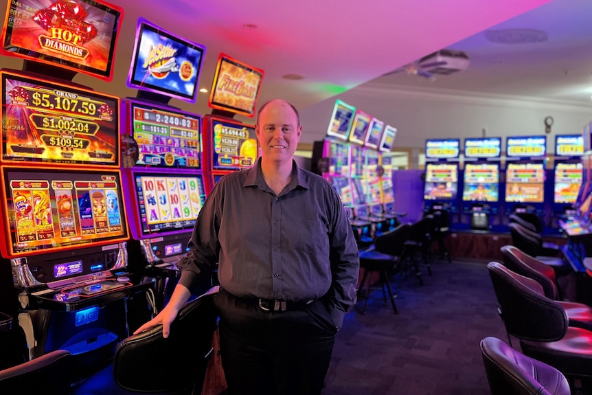 A balding man in a shirt standing in front of a row of poker machines smiling