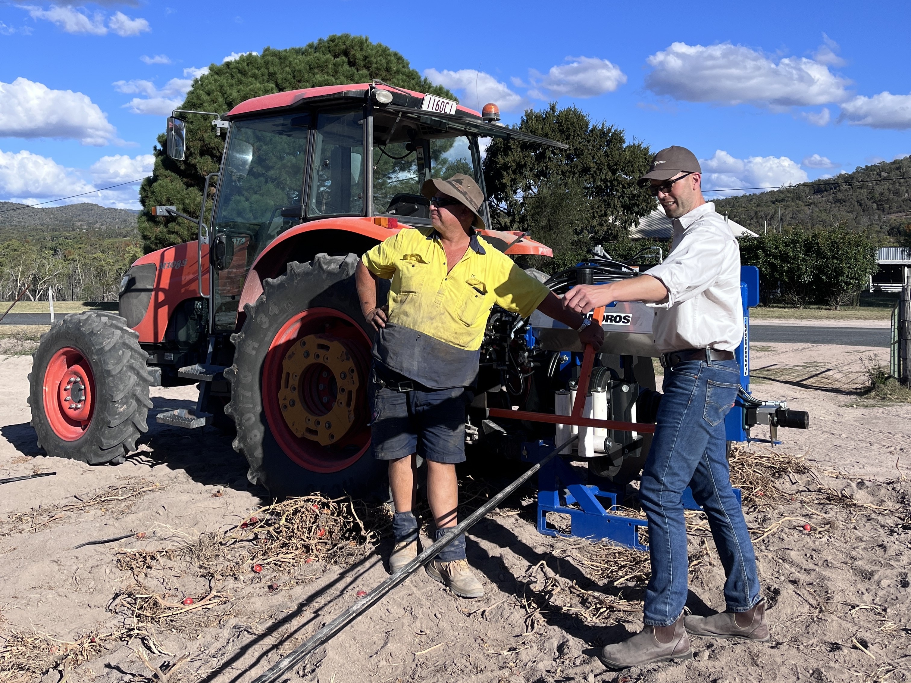 Two men stand near a tractor in a paddock, unrolling a long strip of black plastic.