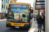 A yellow bus headed for Tranmere stops in the Hobart CBD at a bus shelter, where people are congregated.  