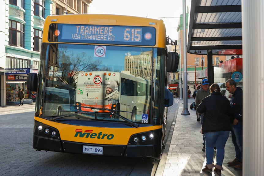 A yellow bus headed for Tranmere stops in the Hobart CBD at a bus shelter, where people are congregated.  