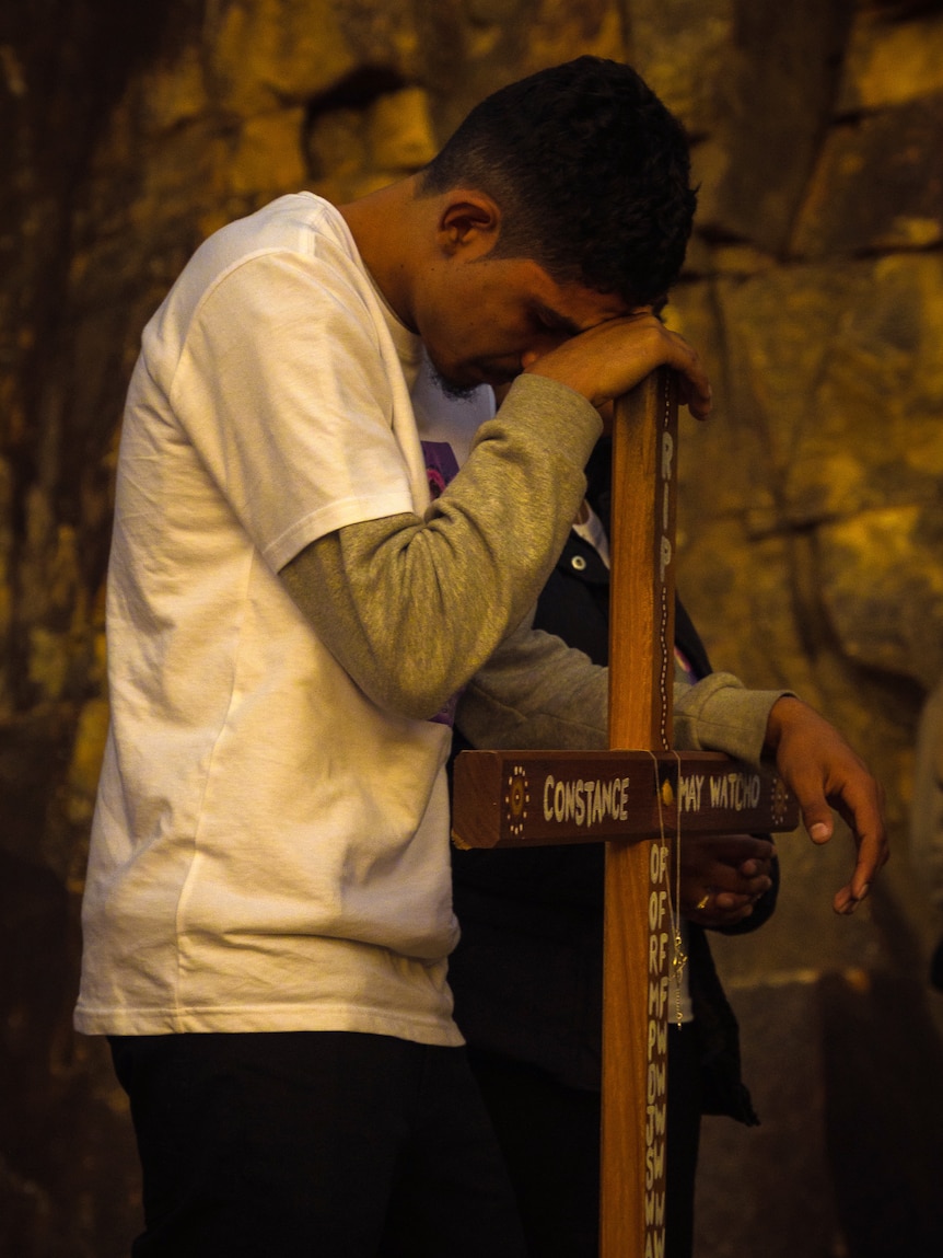 A man leans on a wooden crucifix with cliffs in the background
