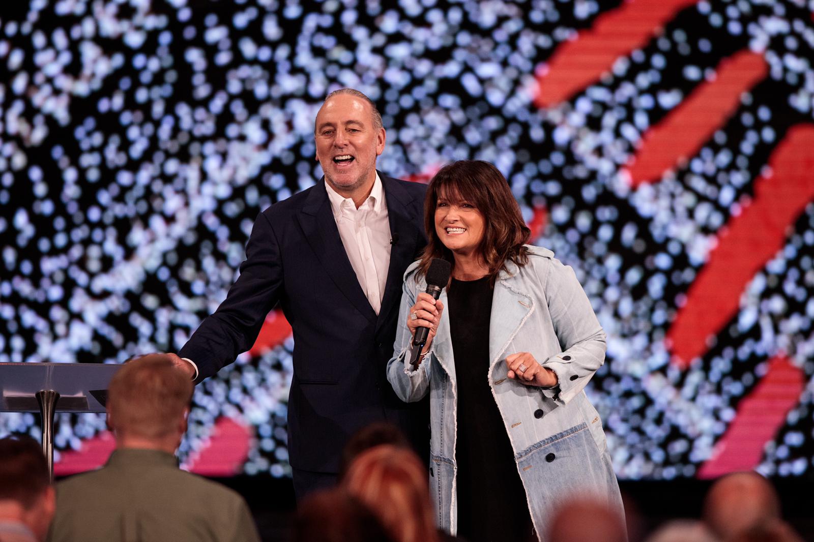 A man in a black suit and white top standing next to a woman in a denim jacket on stage with bright lights