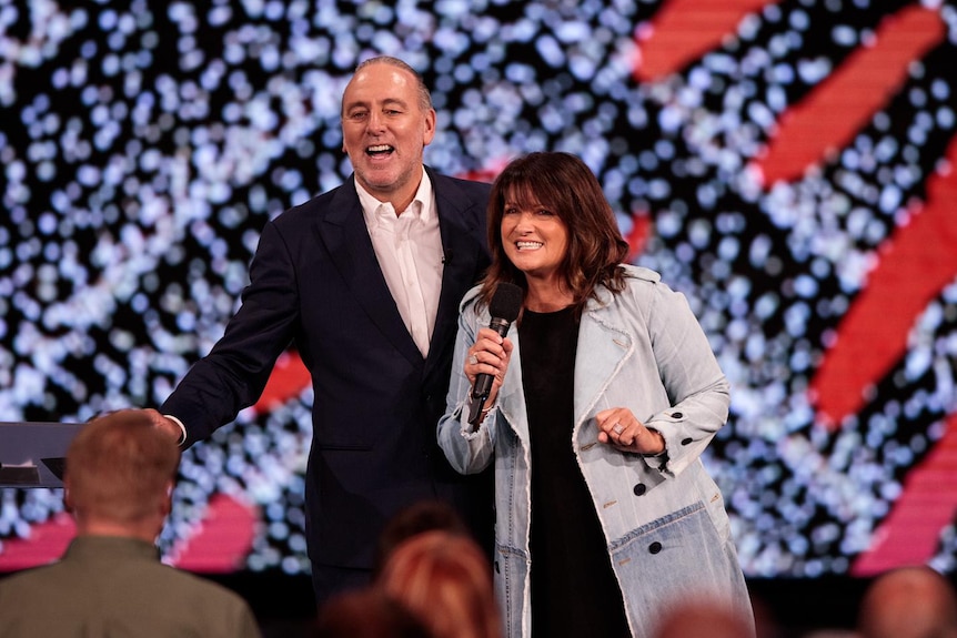 A man in a black suit and white top standing besides a woman in a denim jacket on stage with bright lights