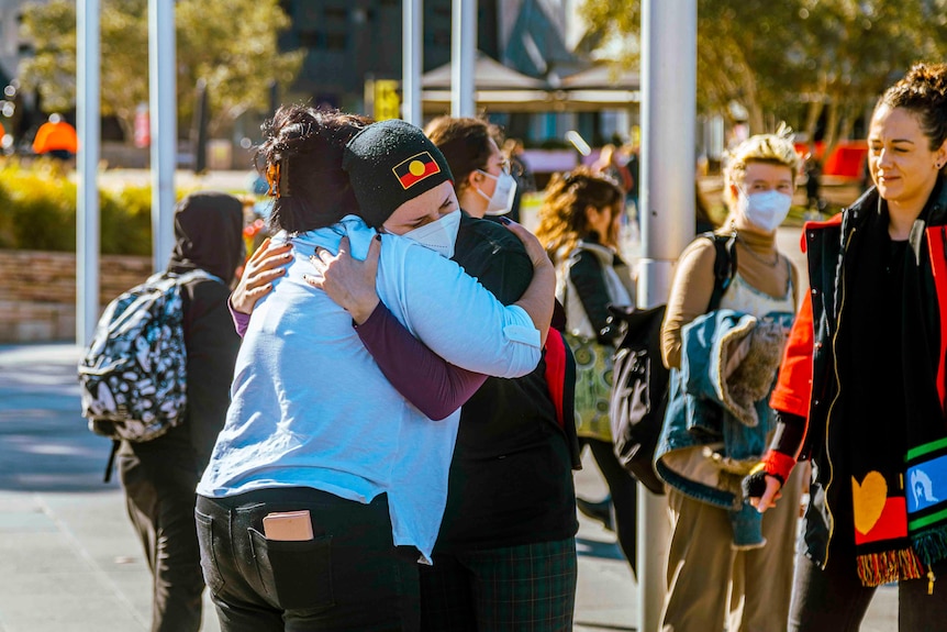 Two women hug, one of them wearing a beanie with the Aboriginal flag, while other community members watch on.