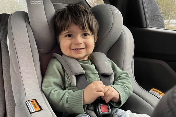A little boy in a green jumper is smiling in a car seat.