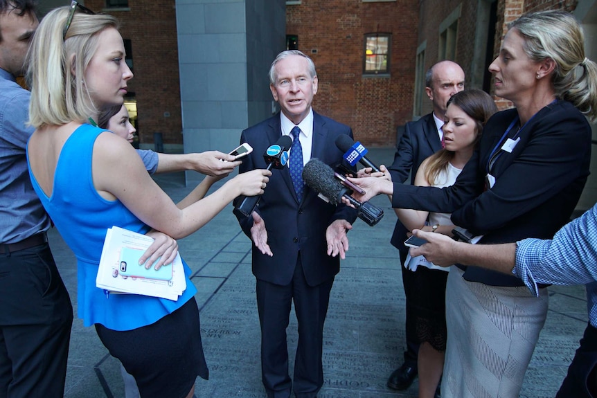 Colin Barnett talks with his arms outstretched while surrounded by reporters holding microphones and phones.
