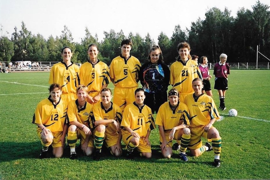 A women's soccer team wearing yellow and green poses for a photo on a pitch