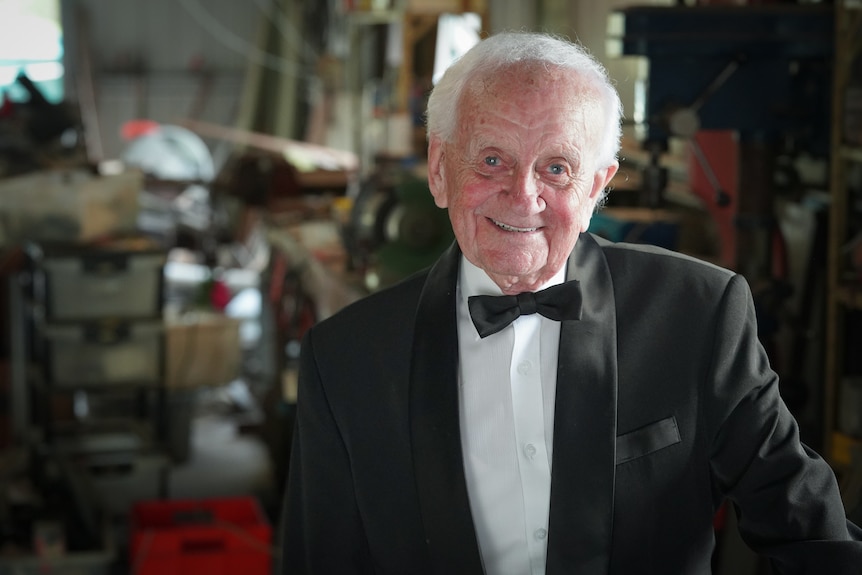 An elderly man in a black suit and bowtie in a workshop/shed