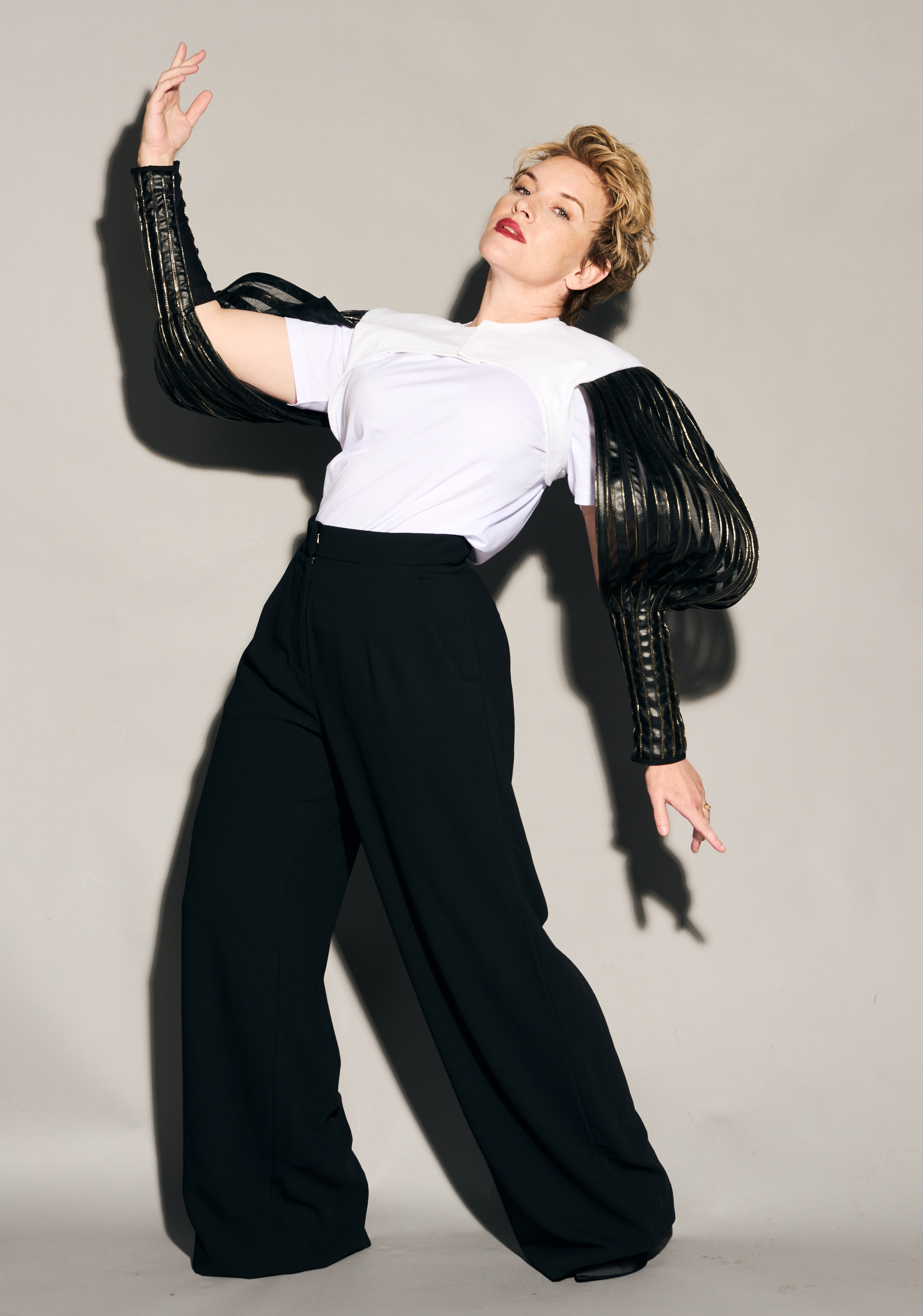 A blonde, short-haired woman in her 40s, leans back, one arm raised. She is wearing black shirt sleeves attached to a white tee.