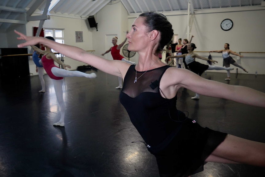 Ballerinas stretch their arms horizontally and raise their left legs as they practice moves in a studio
