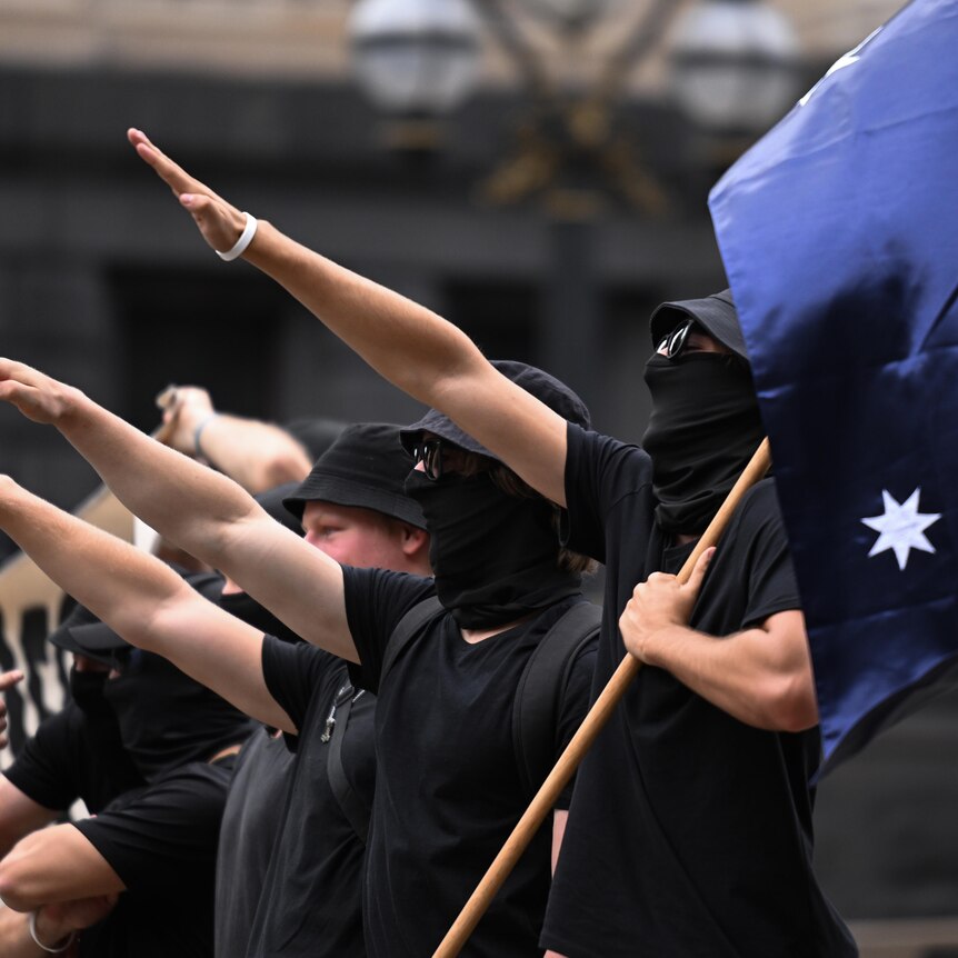 People dressed in black perform a Nazi salute in a line, one of them holding an Australian flag in their non-saluting hand.