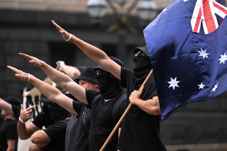 People dressed in black perform a Nazi salute in a line, one of them holding an Australian flag in their non-saluting hand.