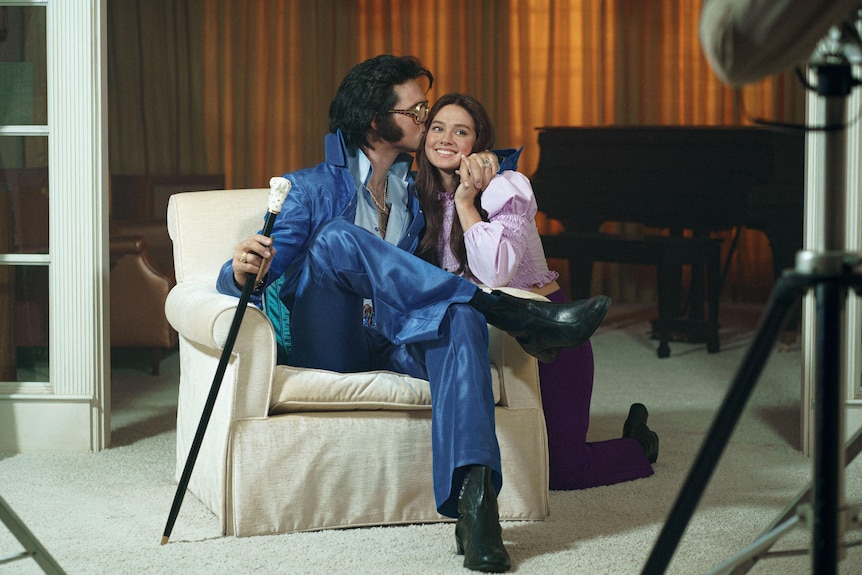 Jacob Elordi in Elvis costume sitting on a lounge as Cailee Spaeny as Priscilla kneels next to him for a kiss in scene from film