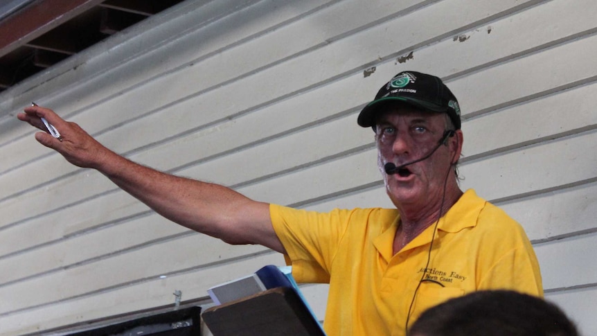 A man in a yellow t-shirt and black cap stands with a microphone and his arm raised outside a weatherboard building.