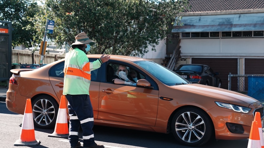 A woman wears a mask and gloves in her orange car as she speaks to a man in high-vis.