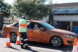 A woman wears a mask and gloves in her orange car as she speaks to a man in high-vis.
