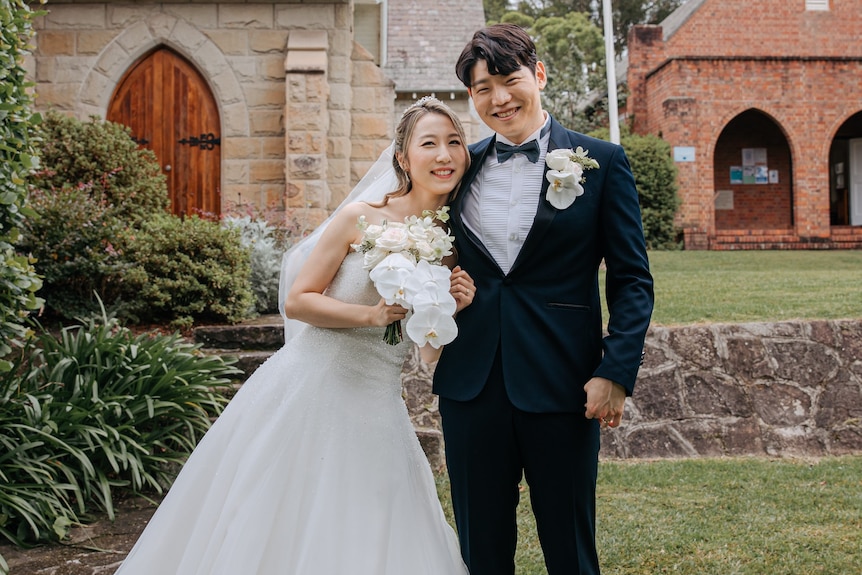 A young Asian couple dressed in wedding outfits smile as they hug each other, outside.