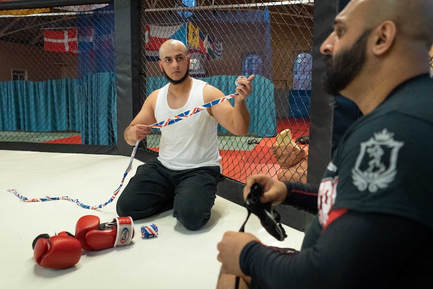 A man unstrapping his hand inside a mixed martial arts ring listens intently to another man.