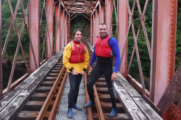 A teenage girl and man stand on a bridge wearing life vests. 