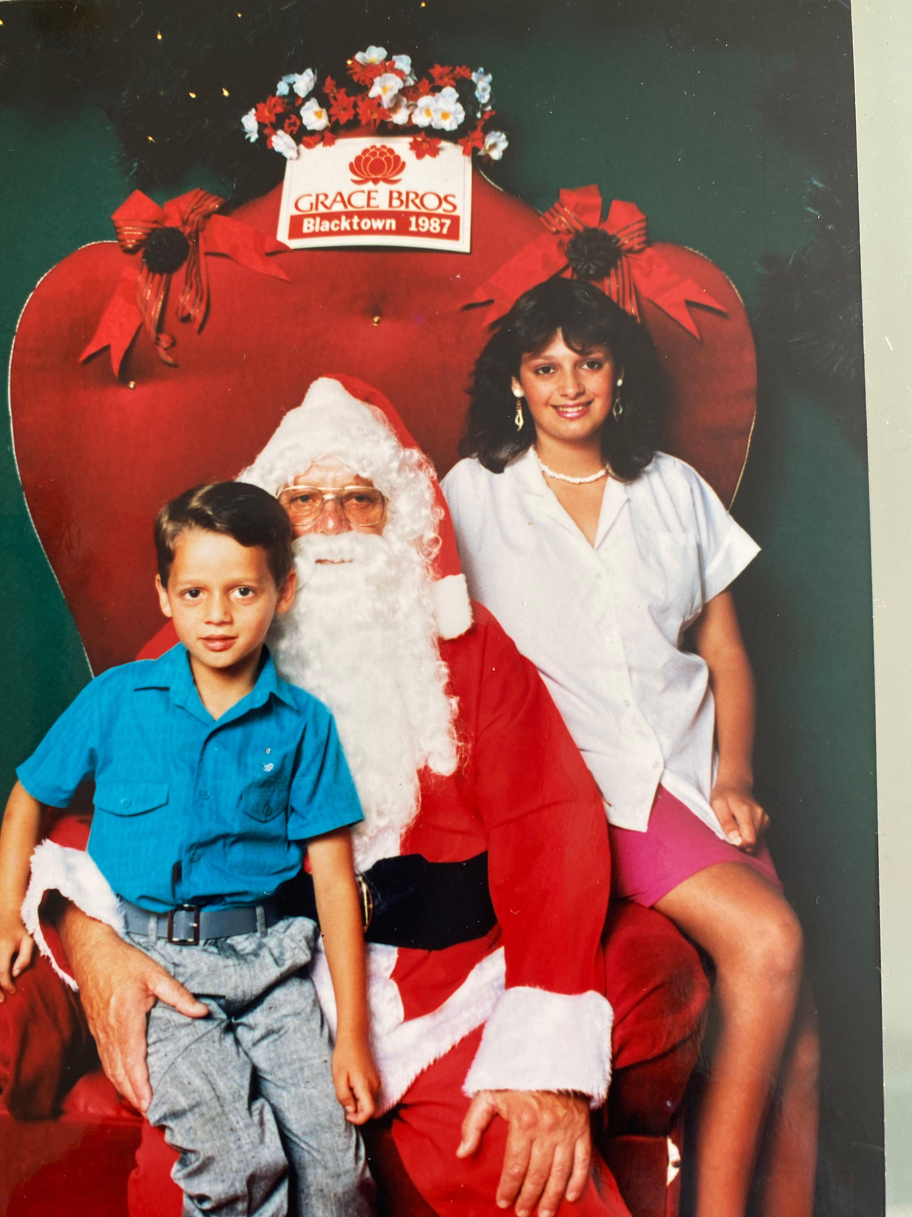A young Maltese Australian boy in a blue shirt sits for a Santa shoot with his older sister who is wearing a white shirt.