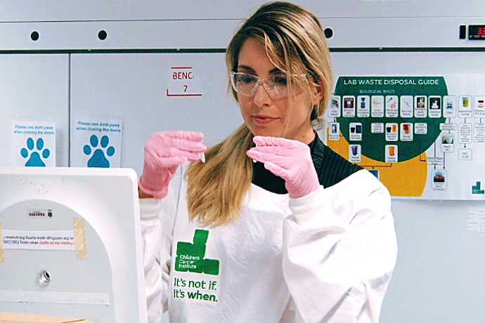 A woman with blonde hair wears a lab coat and looks at viles 