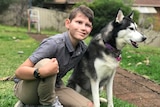 A young boy kneels next to a Husky dog. The boy is smiling, looking just past the camera, while the dog's attention is elsewhere