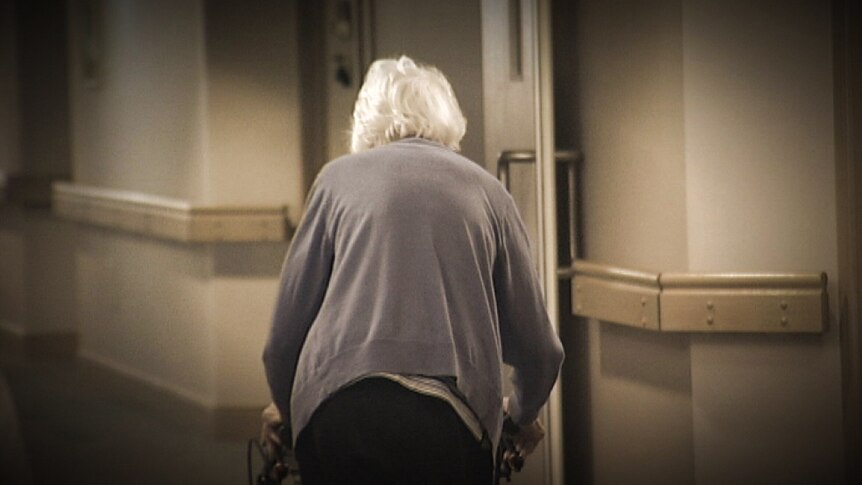 An elderly woman with white hair and a walking frame with her back turned in a corridor.