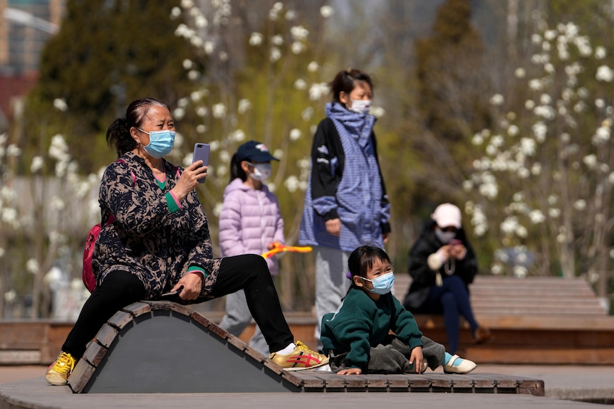 Two women wearing face masks supervise their masked children as they play in a playground in the sunshine.