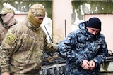 A Ukrainian sailor in camouflage gear is escorted by a Russian intelligence officer, also in camouflage gear, into court.