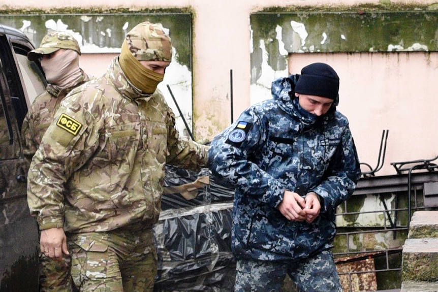 A Ukrainian sailor in camouflage gear is escorted by a Russian intelligence officer, also in camouflage gear, into court.