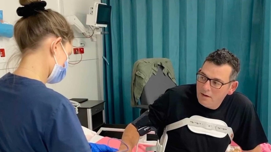 a health worker in a mask stands next to daniel andrews who is sitting on a hospital bed
