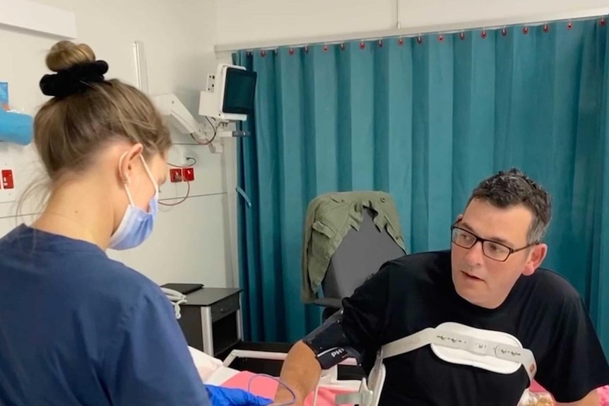 a health worker in a mask stands next to daniel andrews who is sitting on a hospital bed.