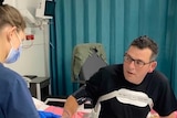 a health worker in a mask stands next to daniel andrews who is sitting on a hospital bed