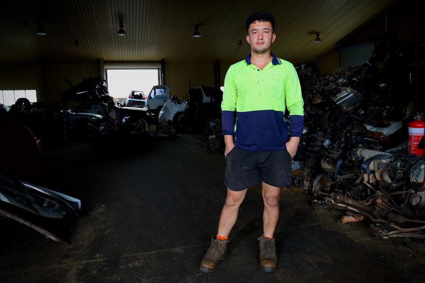 A man in a yellow high-vis shirt, black shorts and shoes standing inside a warehouse of autoparts, looking serious.