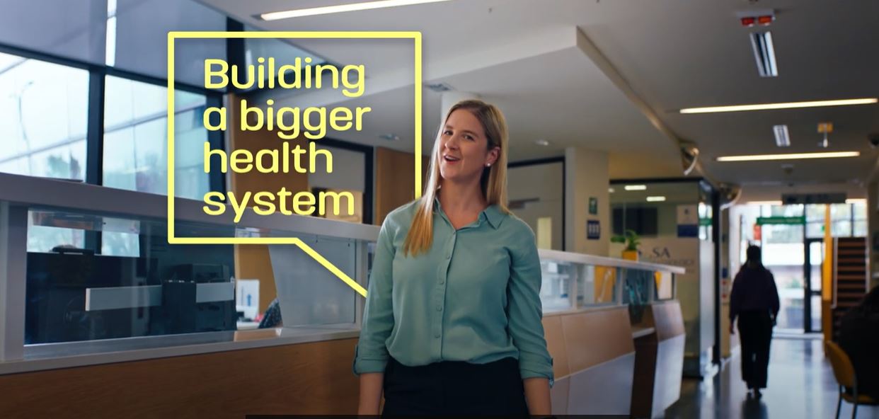 A screenshot of a woman inside a clinic with a graphic behind her &#x27;Building a bigger health system&#x27;