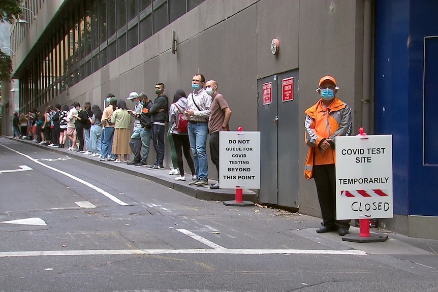 A man in high-vis jacket stands next to a sign saying covid test site temporarily closed as people queue behind him.