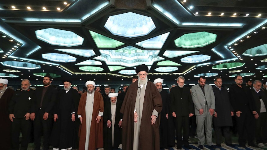 Iran's supreme leader stands with a group of men beneath a star-shaped dome hands in Islamic praying position