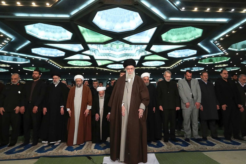 Iran's supreme leader stands with a group of men beneath a star-shaped dome hands in Islamic praying position