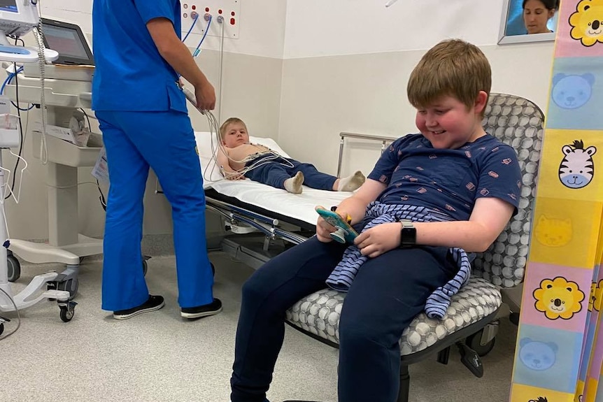 Boy sits in chair while brother is with nurse on hospital bed behind him. 