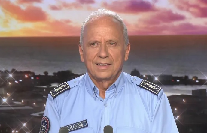 Un hombre de pelo gris y uniforme está sentado en lo que parece ser un estudio de televisión con un puerto como telón de fondo.