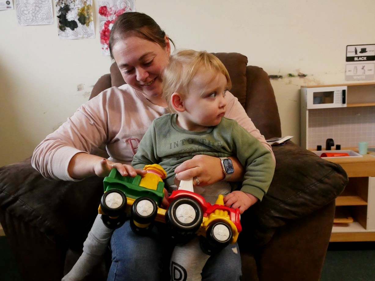 A white woman with her dark hair tied back holds her young male child on her lap and has a toy truck in her hand.