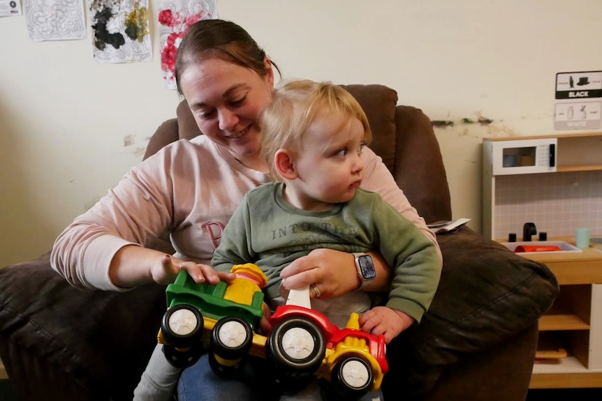A white woman with her dark hair tied back holds her young male child on her lap and has a toy truck in her hand.