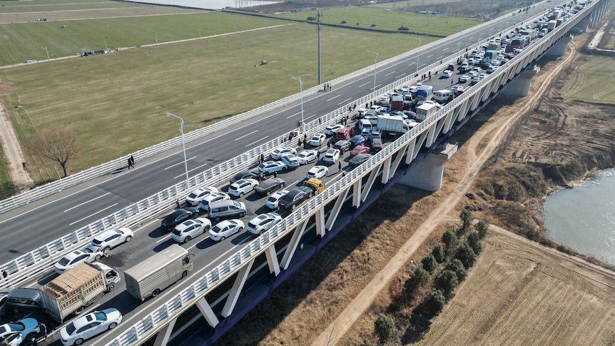 A huge number of cars crashed into each other on a long bridge across a river and paddocks.